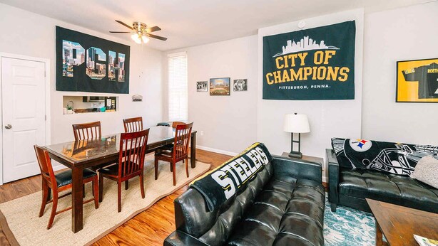 Ein Wohnzimmer mit einer schwarzen Ledersofa, einem Holzesstisch, Stühlen, einem Deckenventilator und Sportmemorabilien an den Wänden.