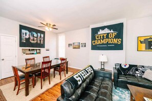 A living room with a black leather couch, wooden dining table, and chairs, a ceiling fan, and sports memorabilia on the walls.