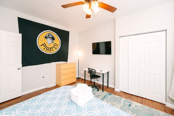 A bedroom with a bed, dresser, desk, chair, TV, wardrobe, and a flag on the wall.