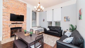 A living room with a brick fireplace, a flat-screen TV, a dining table with chairs, a black leather sofa, and a chandelier. - Spectacular Location - Walkable Entertainment (Pittsburgh)