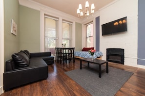 A living room with a black sofa, a coffee table, a dining table with chairs, a fireplace, and a TV mounted on the wall.