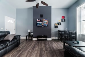 A modern living room with a dark wood floor, a black leather sofa, a dining table with chairs, a ceiling fan, and a mounted TV.