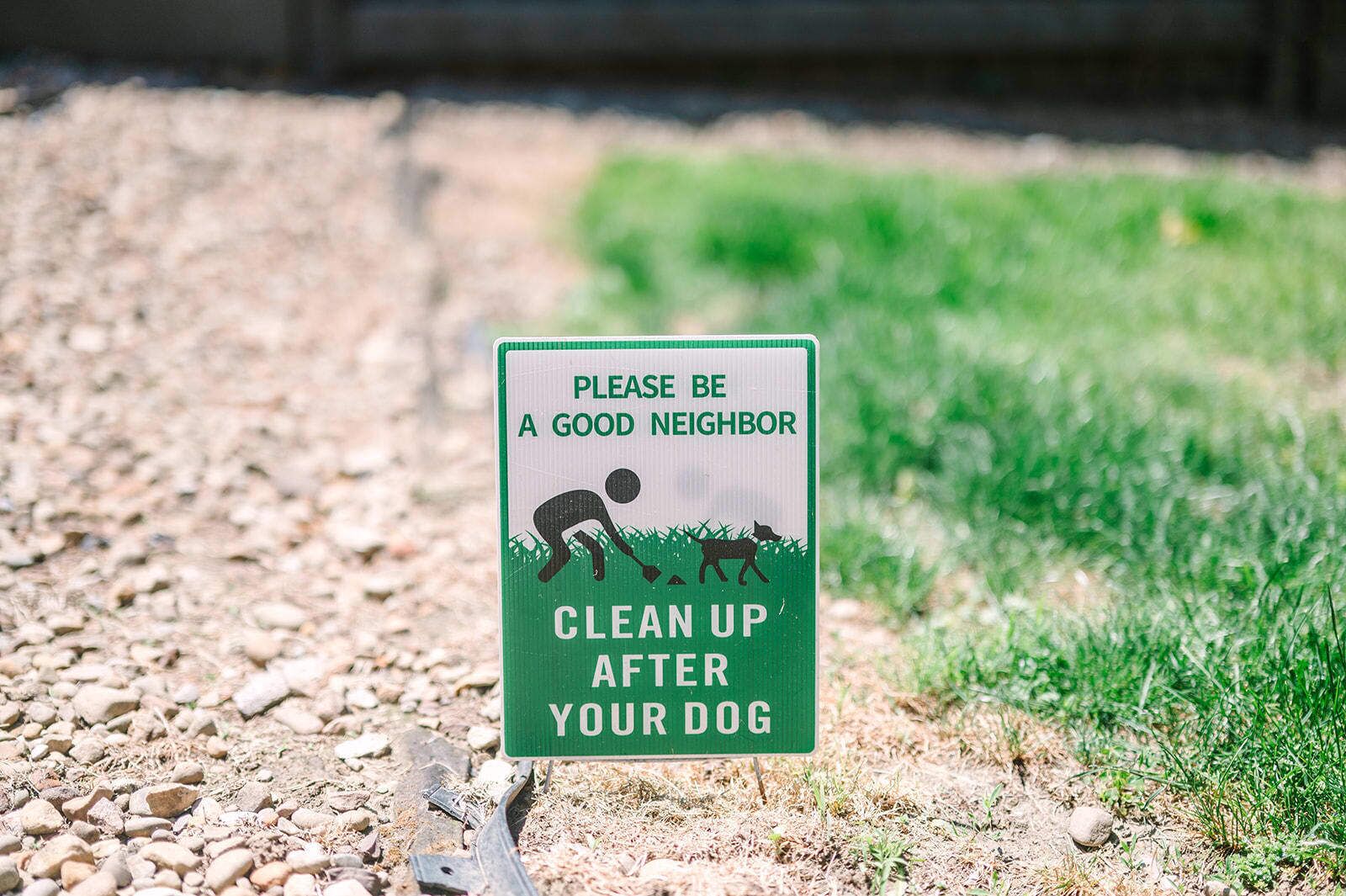 A sign on a gravel path with grass on either side, requesting dog owners to clean up after their pets.