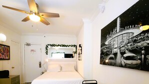 A hotel room with a bed, a desk, a ceiling fan, a painting of a car on the wall, and a door with a sign.