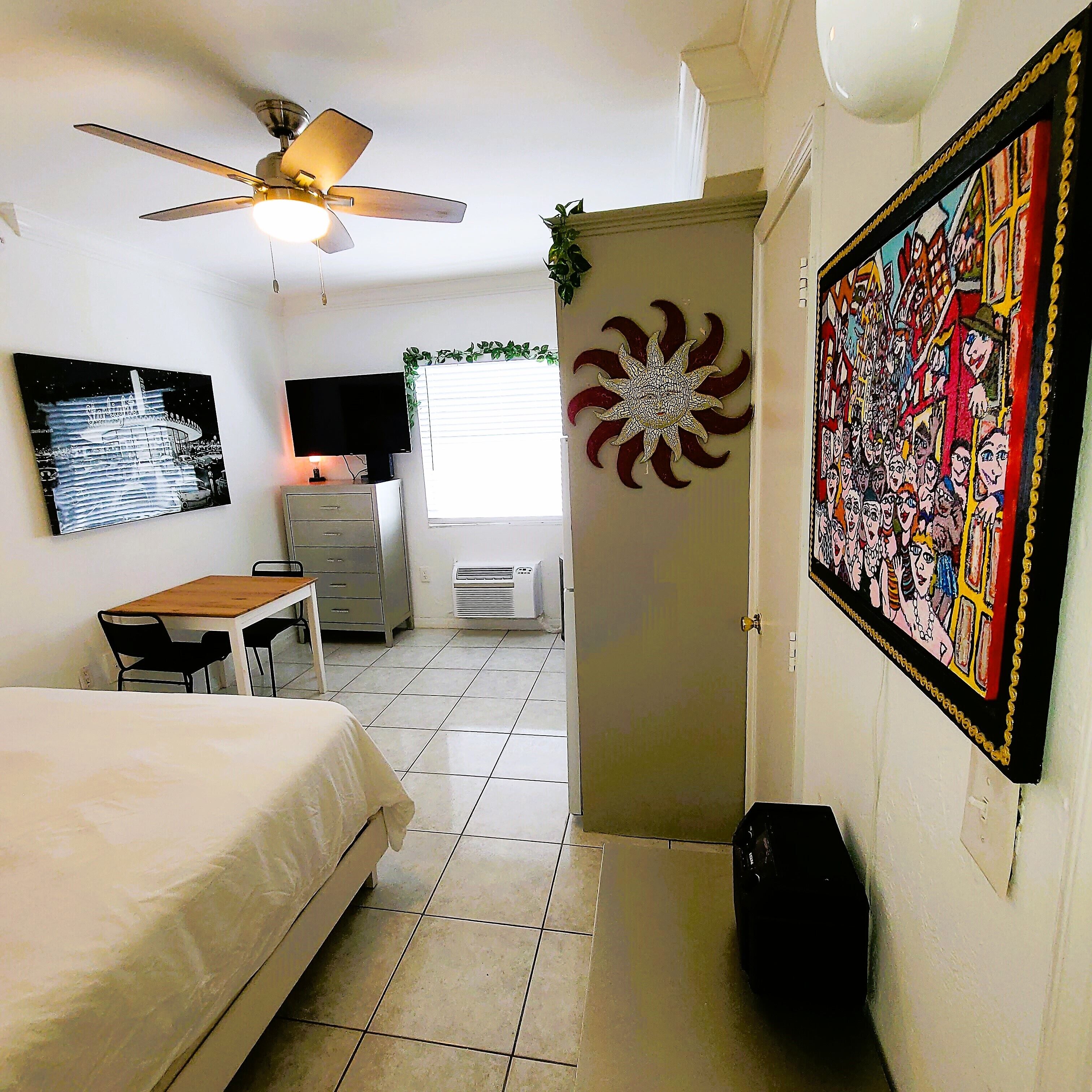 A hotel room with a bed, a desk, a ceiling fan, a TV, a window with blinds, a wall decoration, and a door.