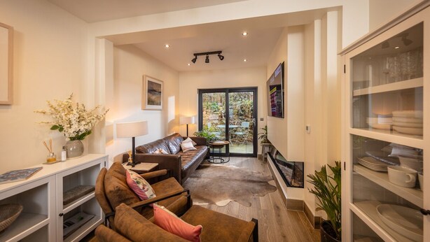 A modern living room with a sofa, coffee table, and a TV. There is a glass door leading to a patio with outdoor furniture. - Downshire Haven (Holywood)