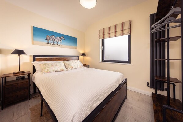 Un dormitorio con cama, dos mesitas de noche, un tocador, una ventana con cortinas, un cuadro en la pared y un armario empotrado.