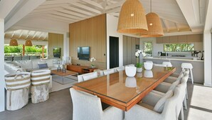 Una sala de estar moderna con una mesa de comedor de vidrio, asientos cómodos, un televisor grande y una cocina con vista a zonas verdes.