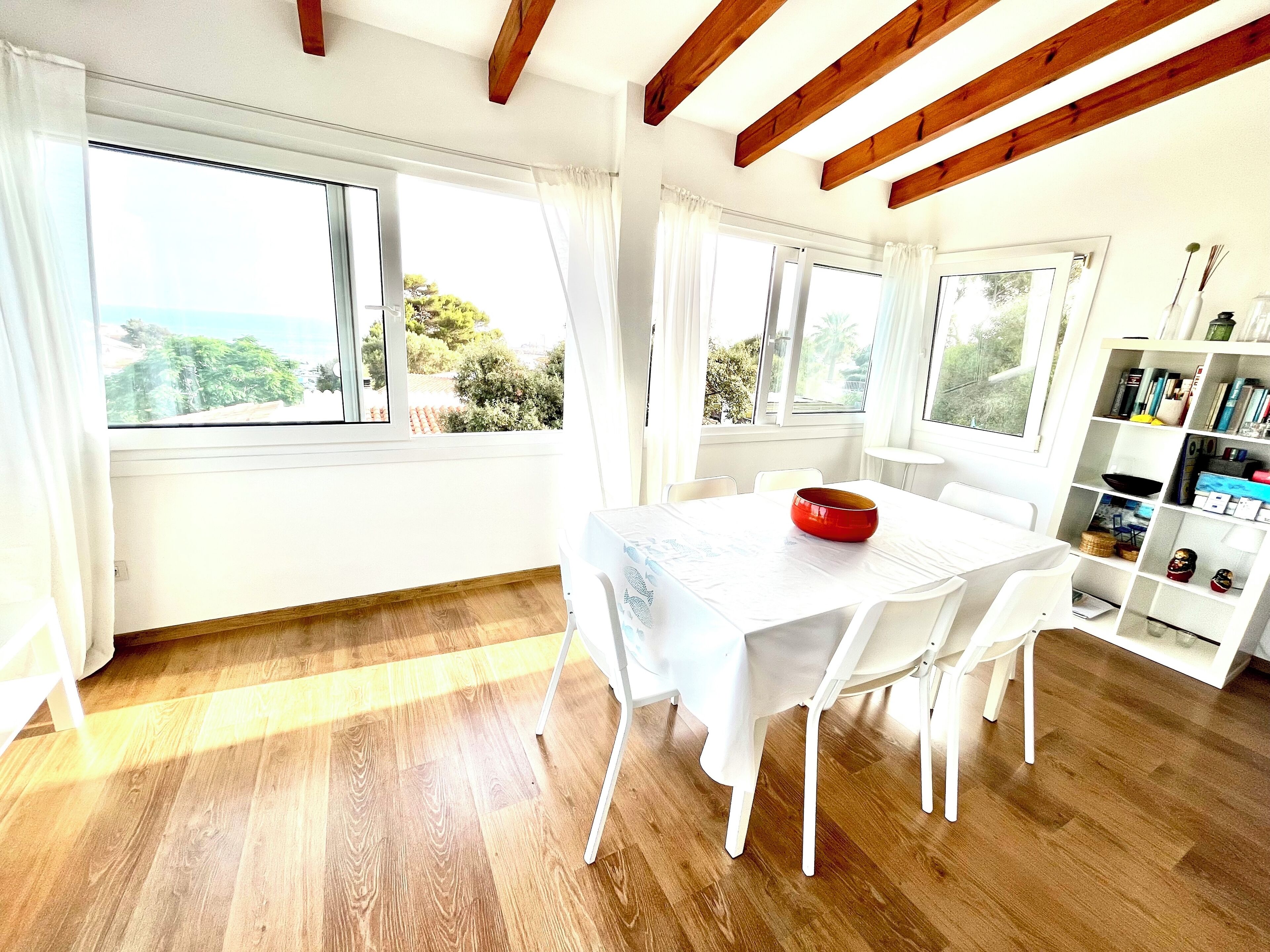 A bright room with wooden beams, a white table with chairs, and a bookshelf.
