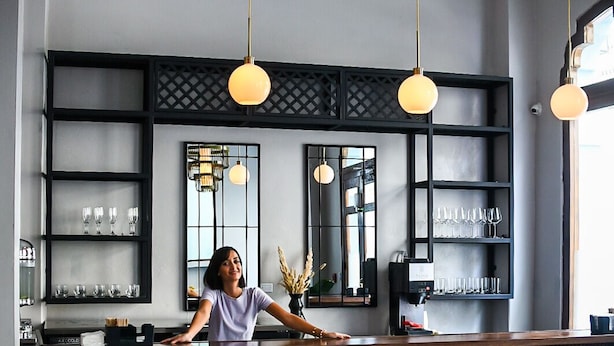 A bar area with a bartender, shelves with glasses, hanging pendant lights, and a wooden counter.