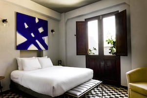 Habitación de hotel con una cama, piso a cuadros, un amplio ventanal con vistas y un cuadro abstracto azul en la pared.