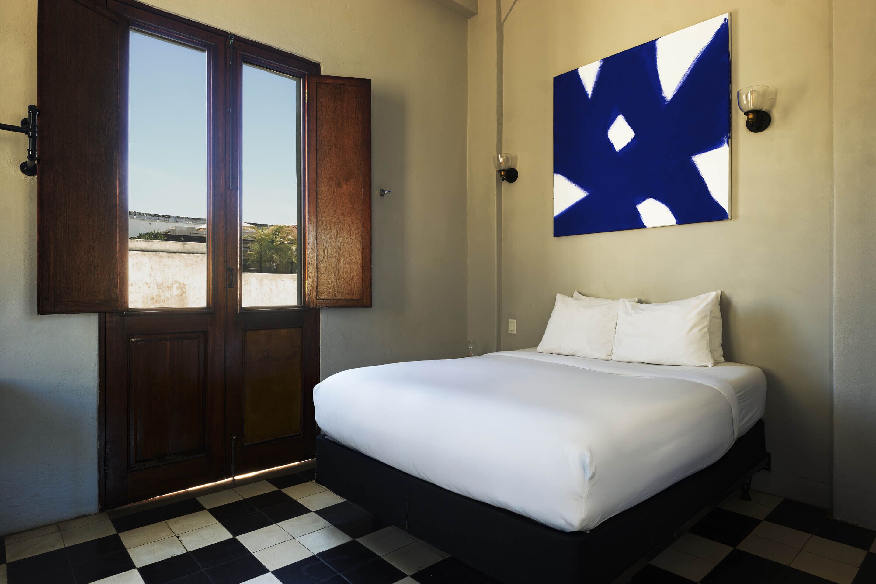 A hotel room with a bed, checkered floor, a large window with a view, and a blue abstract painting on the wall.