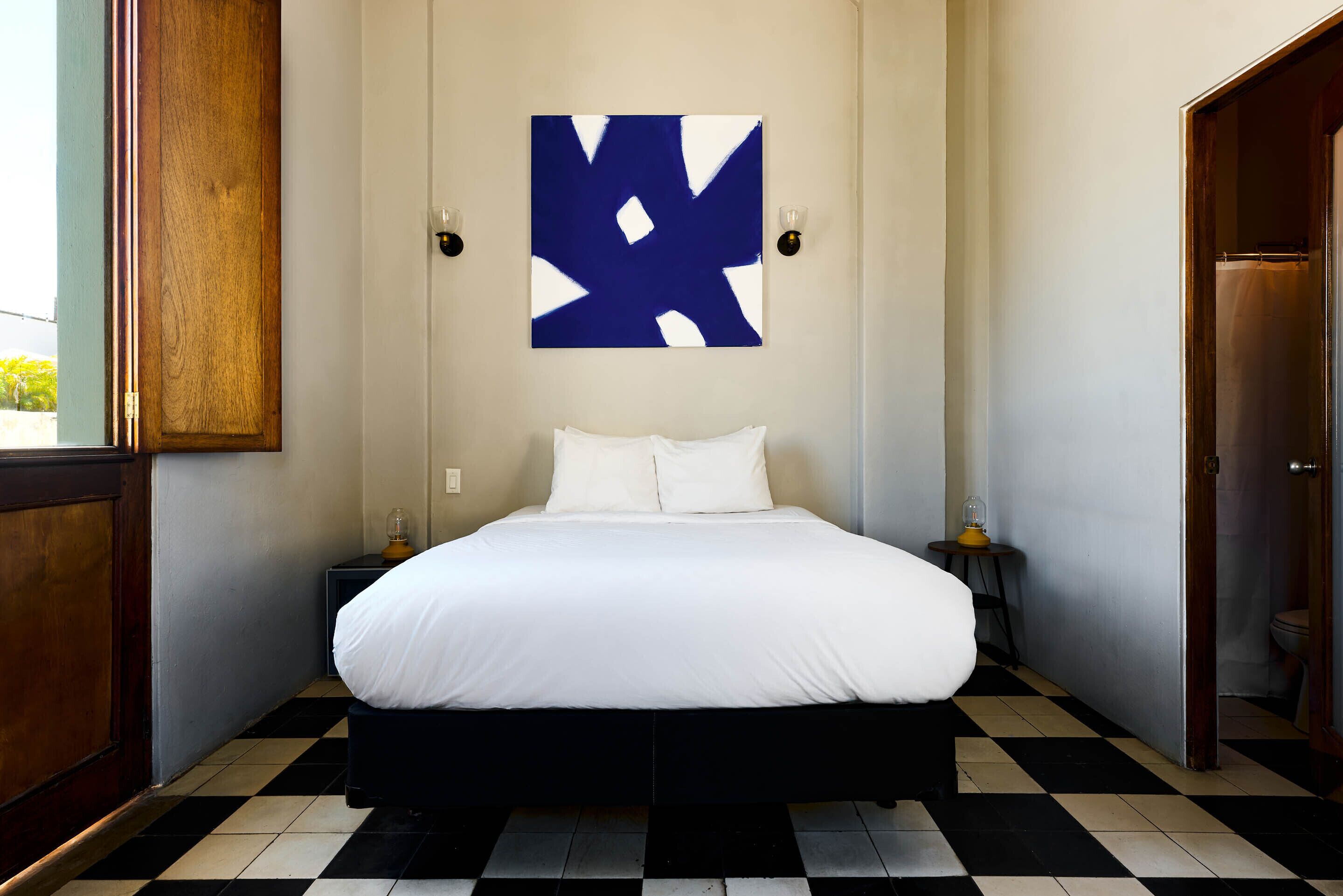 Ein Hotelzimmer mit Schachbrettmuster-Boden, einem Bett mit weißer Bettwäsche, einem blauen abstrakten Gemälde an der Wand und einer Holztür.