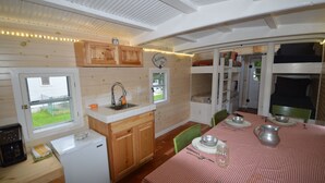 Et kompakt køkken med træskabe, en vask og et lille køleskab. Der er et spiseområde med et bord dækket til to, og en køjeseng i baggrunden.