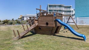 A wooden pirate ship play structure with a slide, surrounded by grass and a white fence.