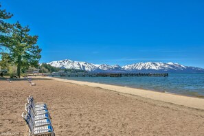 A beach with rows of lounge chairs and a pier extending into a lake with snow-capped mountains in the background.