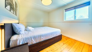 Un dormitorio con suelo de madera, cama con ropa de cama blanca, una lámpara de techo y una ventana con persianas.
