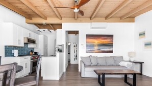 Eine moderne Küche mit weißen Schränken, einer Mikrowelle und einer blauen Spritzschutzwand. Es gibt einen Essbereich mit einem Holztisch und Stühlen, sowie ein Wohnzimmer mit einem grauen Sofa und einem Sonnenuntergangsbild.