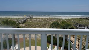 A beach view from a balcony with a wooden deck, chairs, and a boardwalk leading to the beach.
