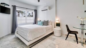 A modern bedroom with a bed, a ceiling fan, an air conditioning unit, a TV, a window with curtains, a bedside table, a chair, and a small table with a plant. - Best Beach Location | Wi-Fi, TV & Amenities (Miami Beach)