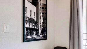 Une pièce moderne avec une table blanche et deux chaises noires, une photographie en noir et blanc encadrée au mur, un luminaire suspendu et une fenêtre avec des rideaux gris.