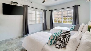 A hotel room with a bed, a ceiling fan, a flat-screen TV mounted on the wall, and a window with curtains. - Best Beach Location | Wi-Fi, TV & Amenities (Miami Beach)