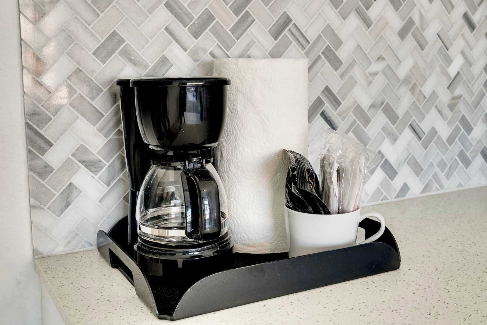 A black coffee maker on a black tray with a white mug and a roll of paper towels.