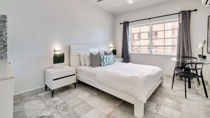 A bedroom with a bed, nightstands, a ceiling fan, a window with curtains, and a small table with chairs. - Best Beach Location | Wi-Fi, TV & Amenities (Miami Beach)
