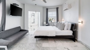 A modern bedroom with a bed, grey sofa, ceiling fan, TV, window, and air conditioning unit. - Best Location SOBE Top Floor 2 min to beach & fun (Miami Beach)