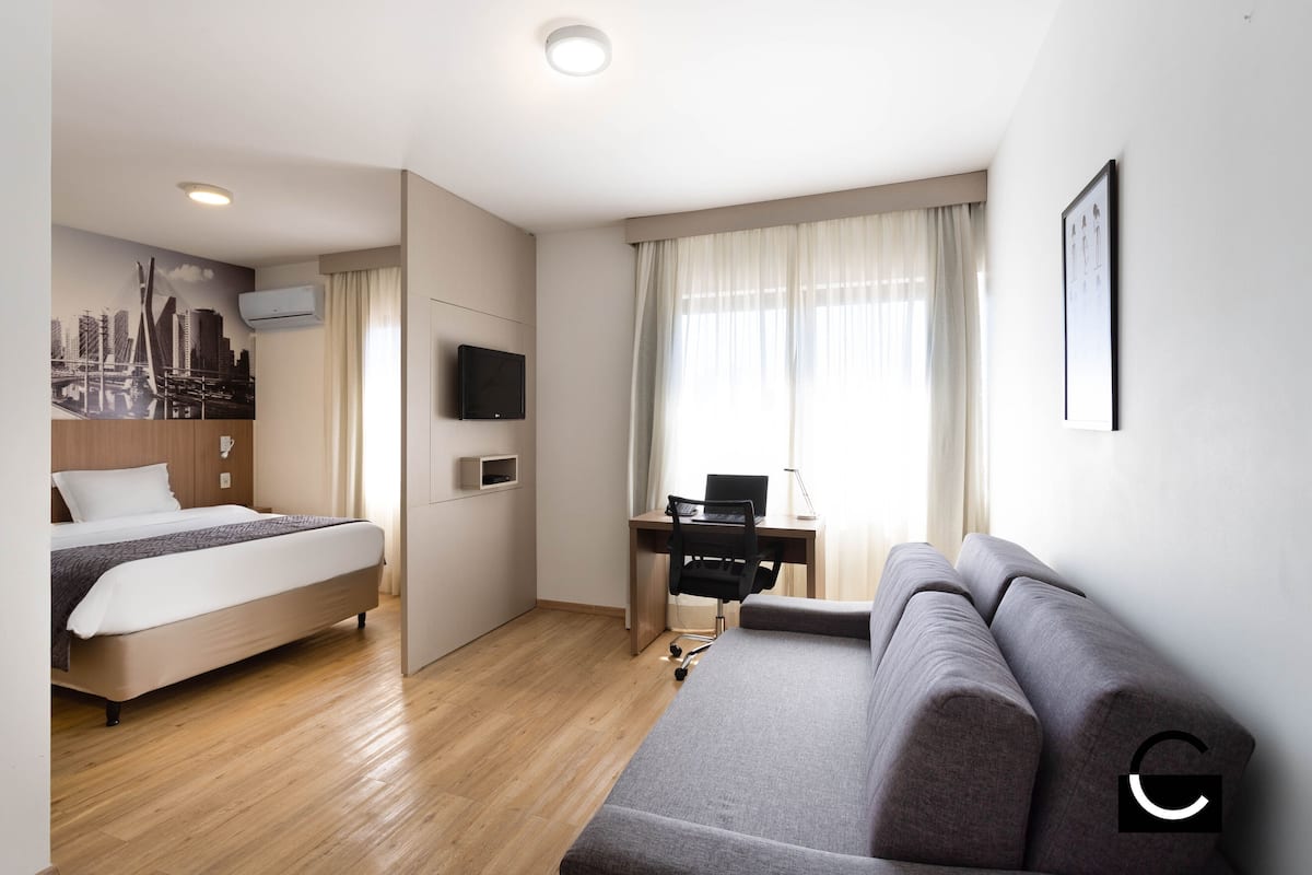 A modern hotel room with a bed, a desk with a chair, a sofa, a TV, and a cityscape mural on the wall.