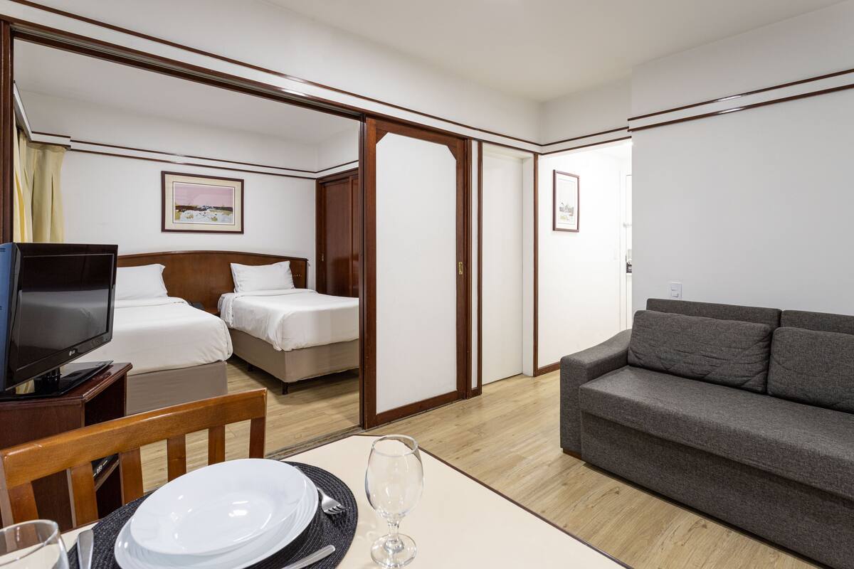 A hotel room with a television, two beds, a sofa, a dining table with plates and cutlery, and a picture on the wall.
