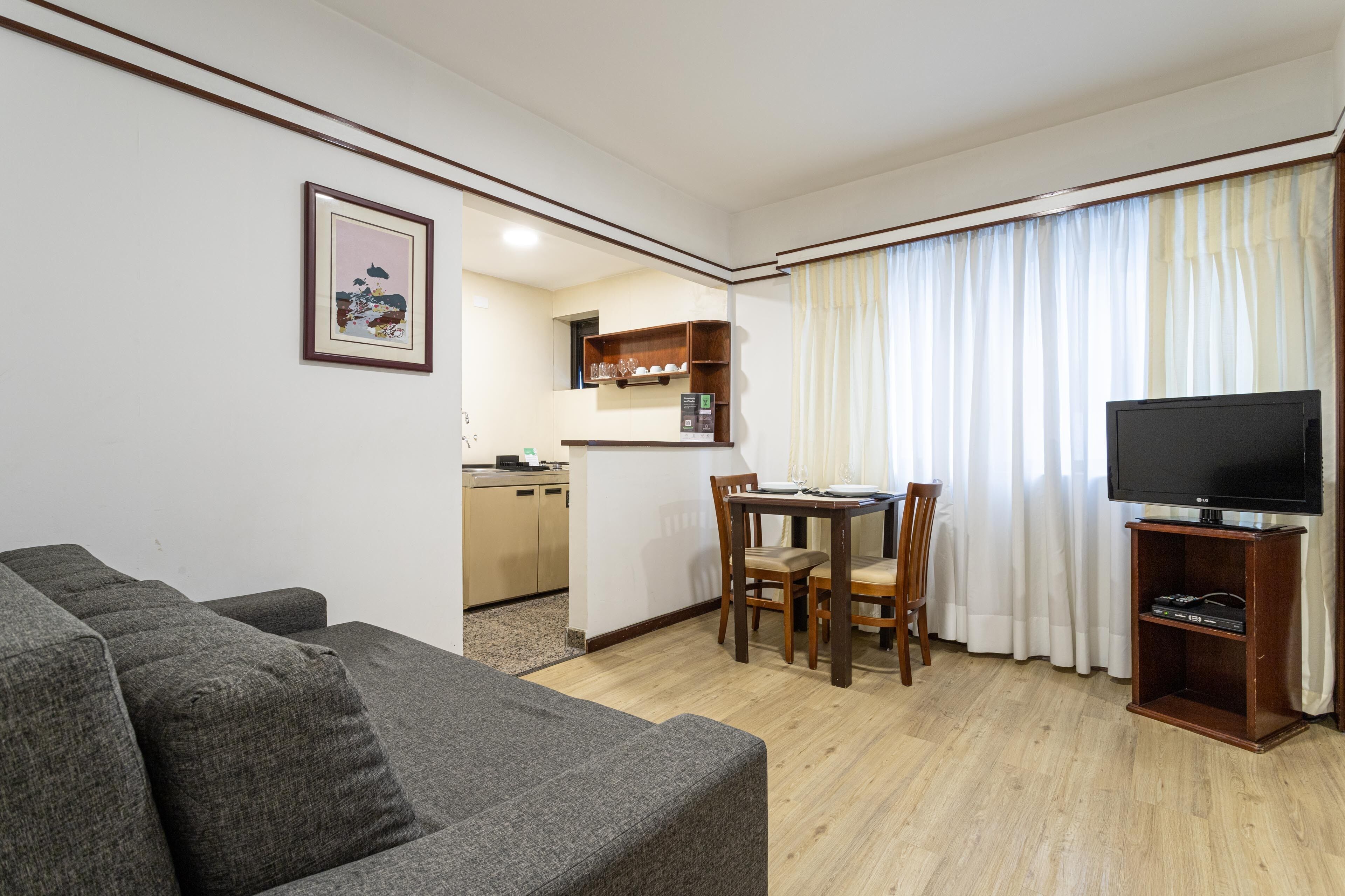 Uma sala de estar com um sofá cinzento, uma mesa de jantar com cadeiras, uma área de cozinha e uma TV sobre um suporte.