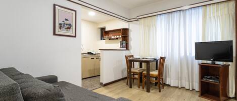 Uma sala de estar com um sofá cinzento, uma mesa de jantar com cadeiras, uma área de cozinha e uma TV sobre um suporte.