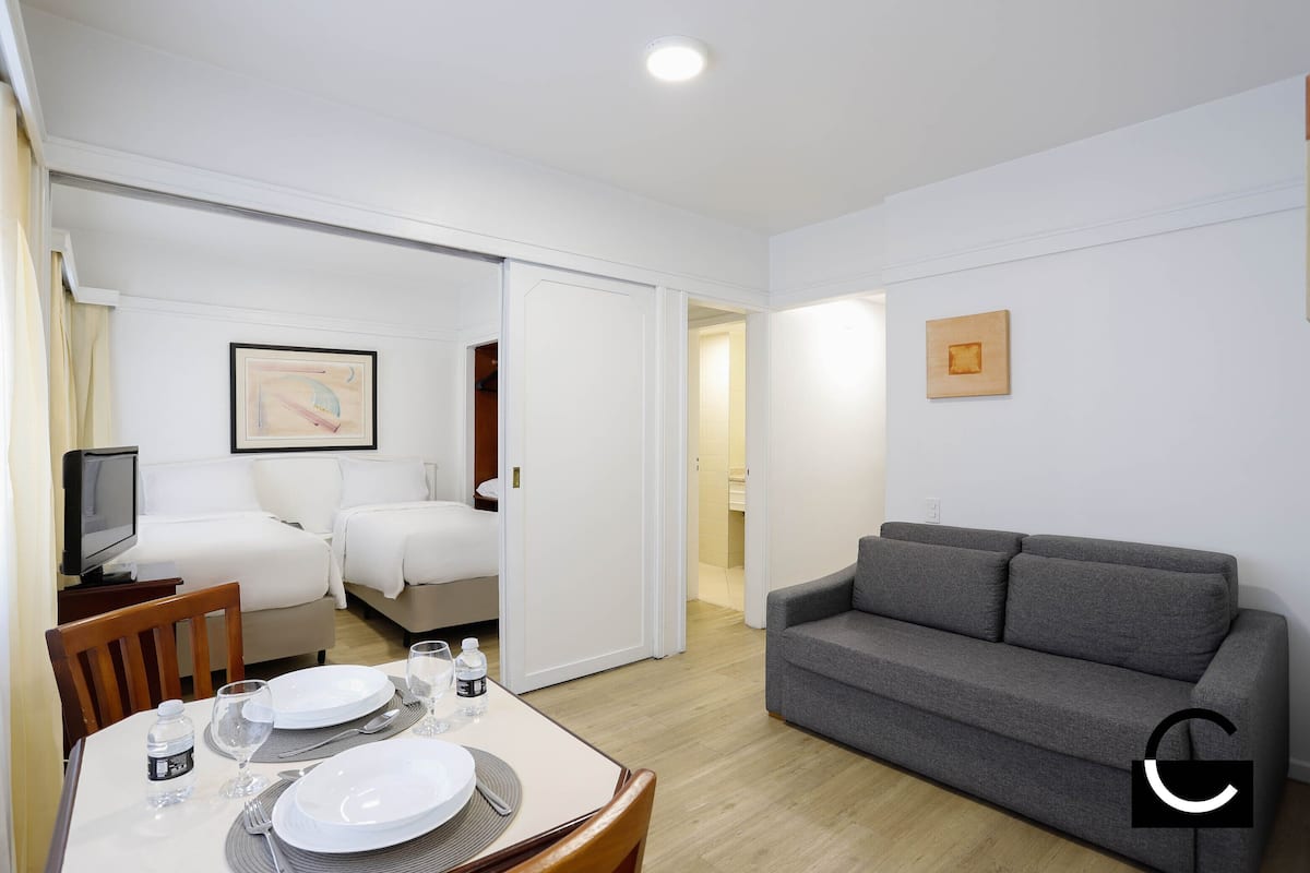 A hotel room with a dining table set for two, a grey sofa, a TV, and a bed.