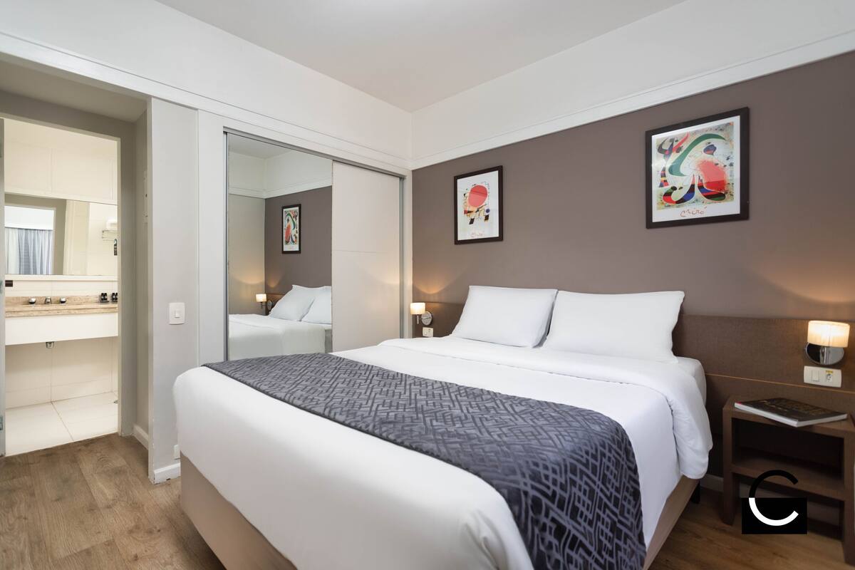 A hotel room with a large bed, a bedside table with a lamp, a mirror, and artwork on the wall.