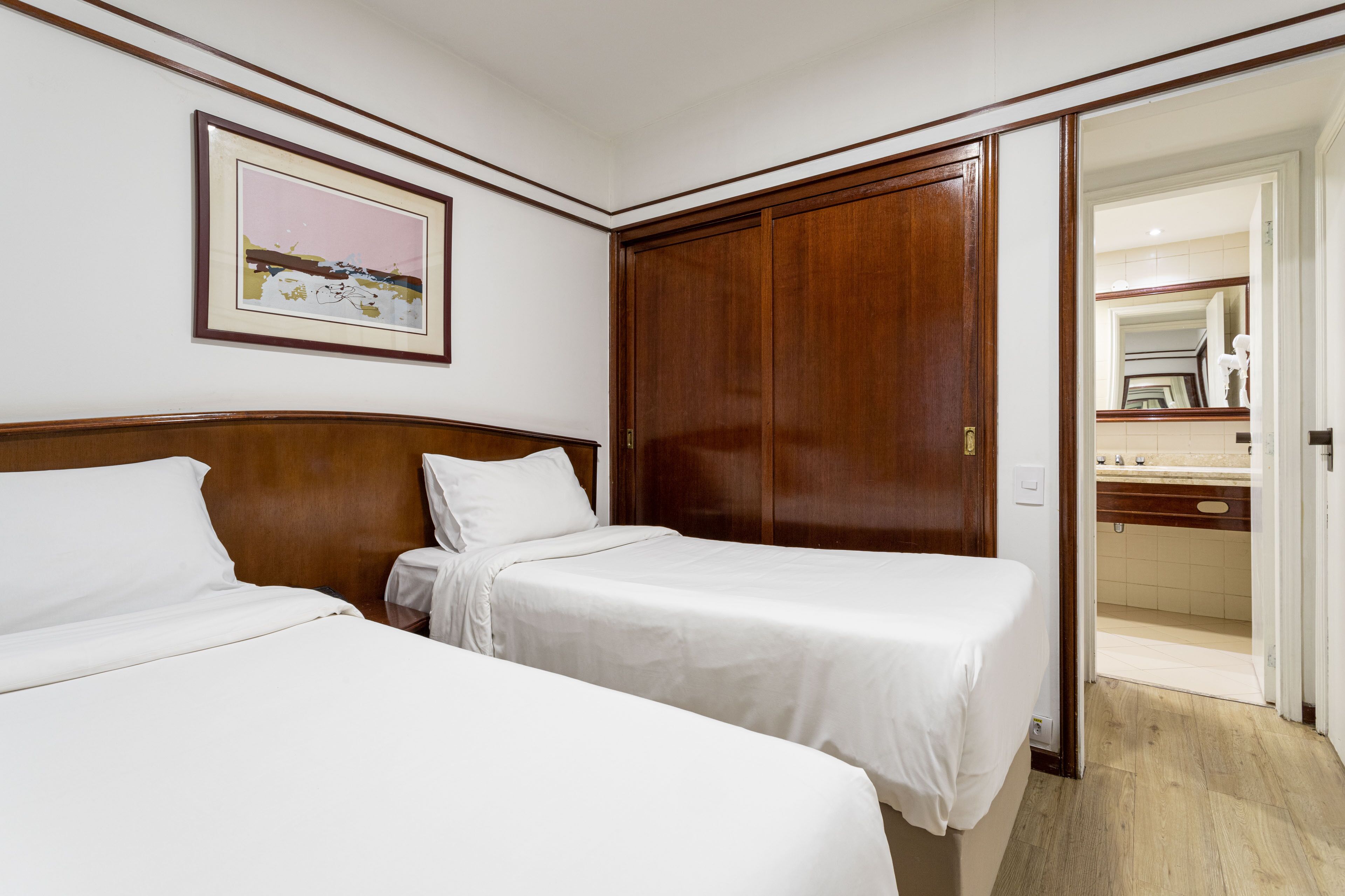 Een hotelkamer met twee éénpersoonsbedden, een houten garderobe, een ingelijst schilderij aan de muur en een badkamer die door een geopende deur zichtbaar is.
