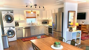 Eine moderne Küche mit Edelstahlgeräten, ein Essbereich mit Holztisch und ein Wohnbereich mit Fernseher und Bücherregal.
