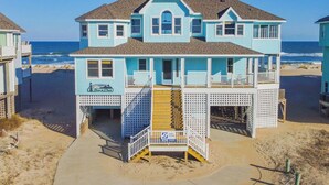 Uma casa de praia de dois andares com varanda, escada que leva à entrada e placa indicando frequência de som do surf.