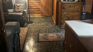 A cozy log cabin interior with a stone wall, a wooden door, a chair with a butterfly pillow, a desk with a lamp, and a small rug.