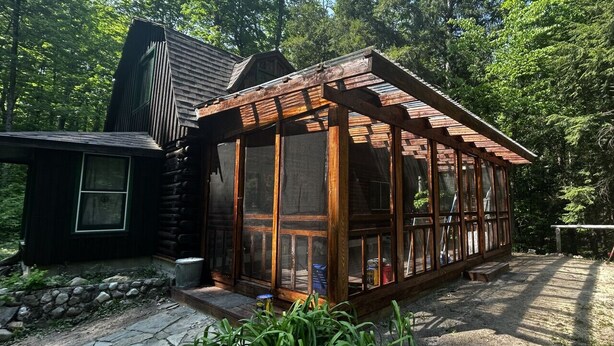 A rustic cabin with a wooden porch and large windows, surrounded by dense forest.