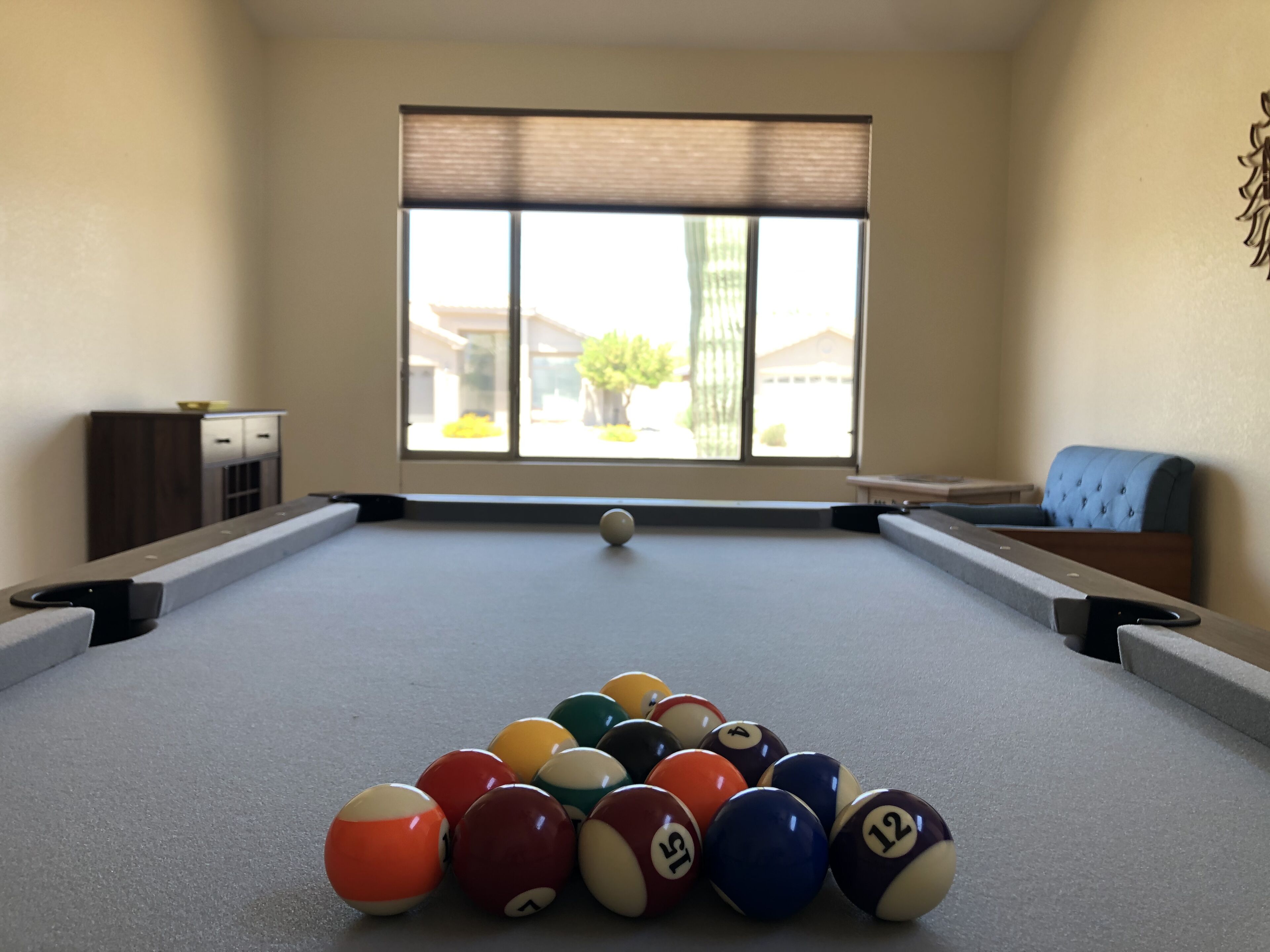 Une table de billard avec les billes disposées en forme de triangle, une fenêtre avec des stores et une vue sur les bâtiments à l’extérieur.
