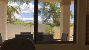 Ein Zimmer mit großem Fenster, das auf eine Terrasse mit Gartenmöbeln und eine Aussicht auf Berge und Palmen blickt.