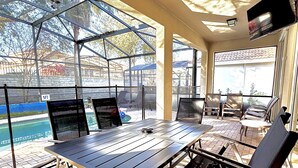 An outdoor patio area with a table and chairs, a pool in the background, and a covered seating area. - 4BR Disney | Pool&Spa | Windsor Hills | Arcade Style Gameroom (Kissimmee)