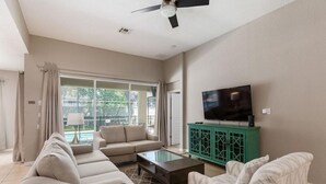A spacious living room with a large window, a ceiling fan, a flat-screen TV, and a green cabinet. - 4BR Disney | Pool&Spa | Windsor Hills | Arcade Style Gameroom (Kissimmee)