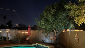 A swimming pool with a tiled edge, surrounded by a fence, and a tree with illuminated lights.