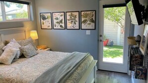 A bedroom with a bed, a nightstand, a lamp, a door leading outside, and four framed botanical prints on the wall.