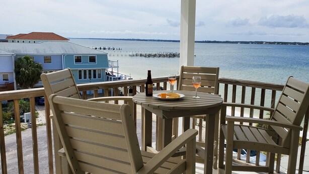 A balcony with wooden chairs and a table, overlooking a calm sea and a pier.
