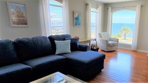 A living room with a dark-colored sectional sofa, a white armchair, and a glass coffee table. There is a view of the sea through large windows and a cabinet with a 'welcome' sign.