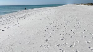 A sandy beach with footprints leading to the water, a clear sky, and a person walking in the distance.