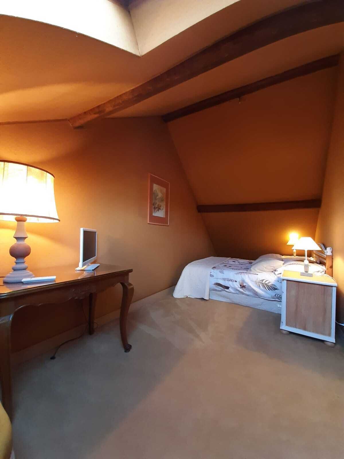 Ein gemütliches Schlafzimmer im Dachgeschoss mit schrägen Wänden, einem Schreibtisch mit Lampe, einem Bett mit Nachttisch und einem gerahmten Bild an der Wand.